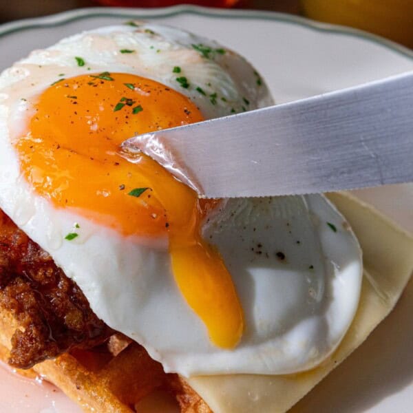 The image features a knife slicing through a runny sunny-side-up egg placed on top of a stack consisting of waffles, fried chicken, and cheese on a white plate.