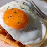 A plate holds a waffle topped with melted cheese, sausage, a sunny-side-up egg, chopped herbs, and a fork on the side.