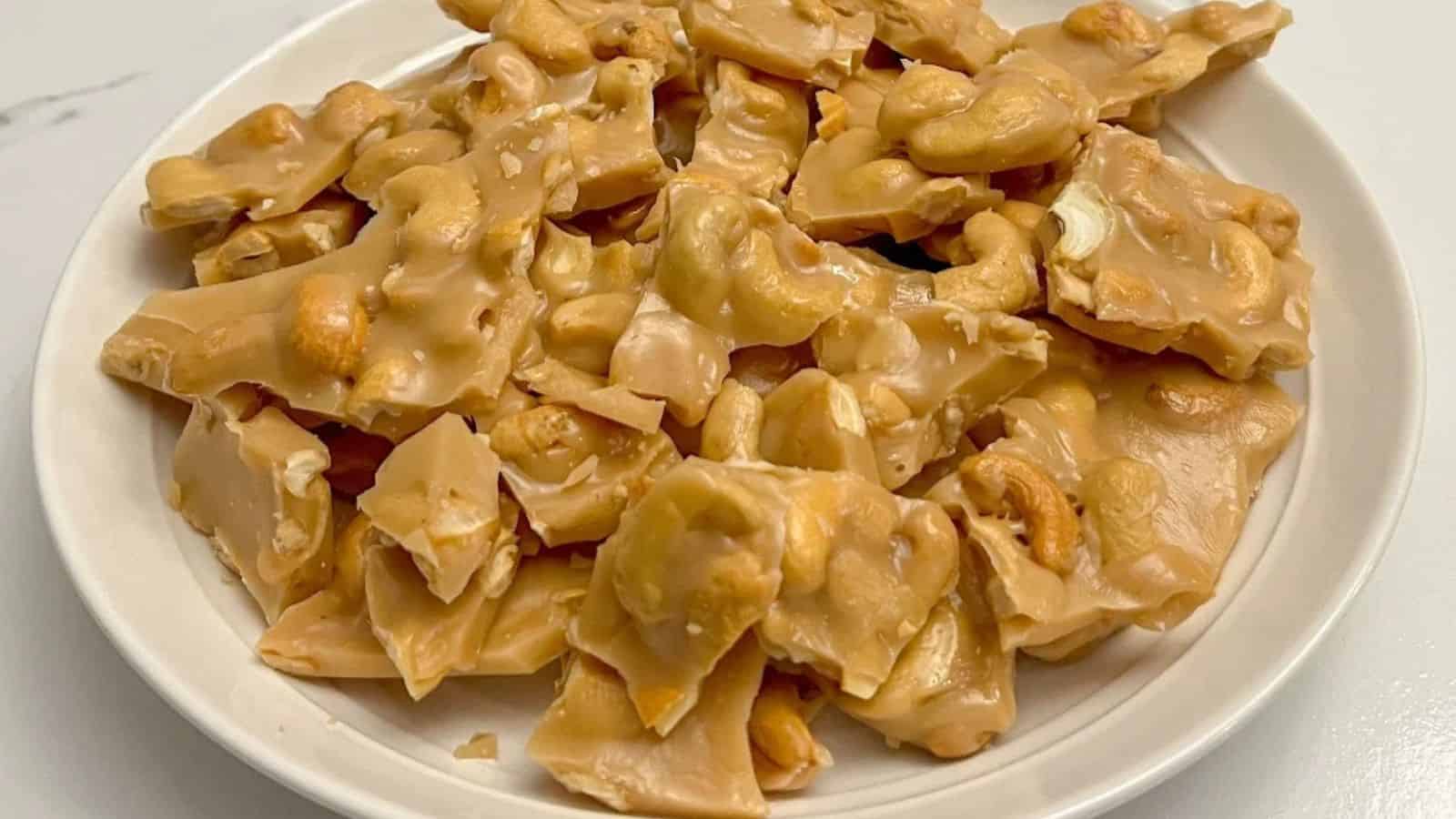 A close-up of a white plate filled with crunchy cashew brittle, showcasing broken, caramel-coated pieces with visible nuts.