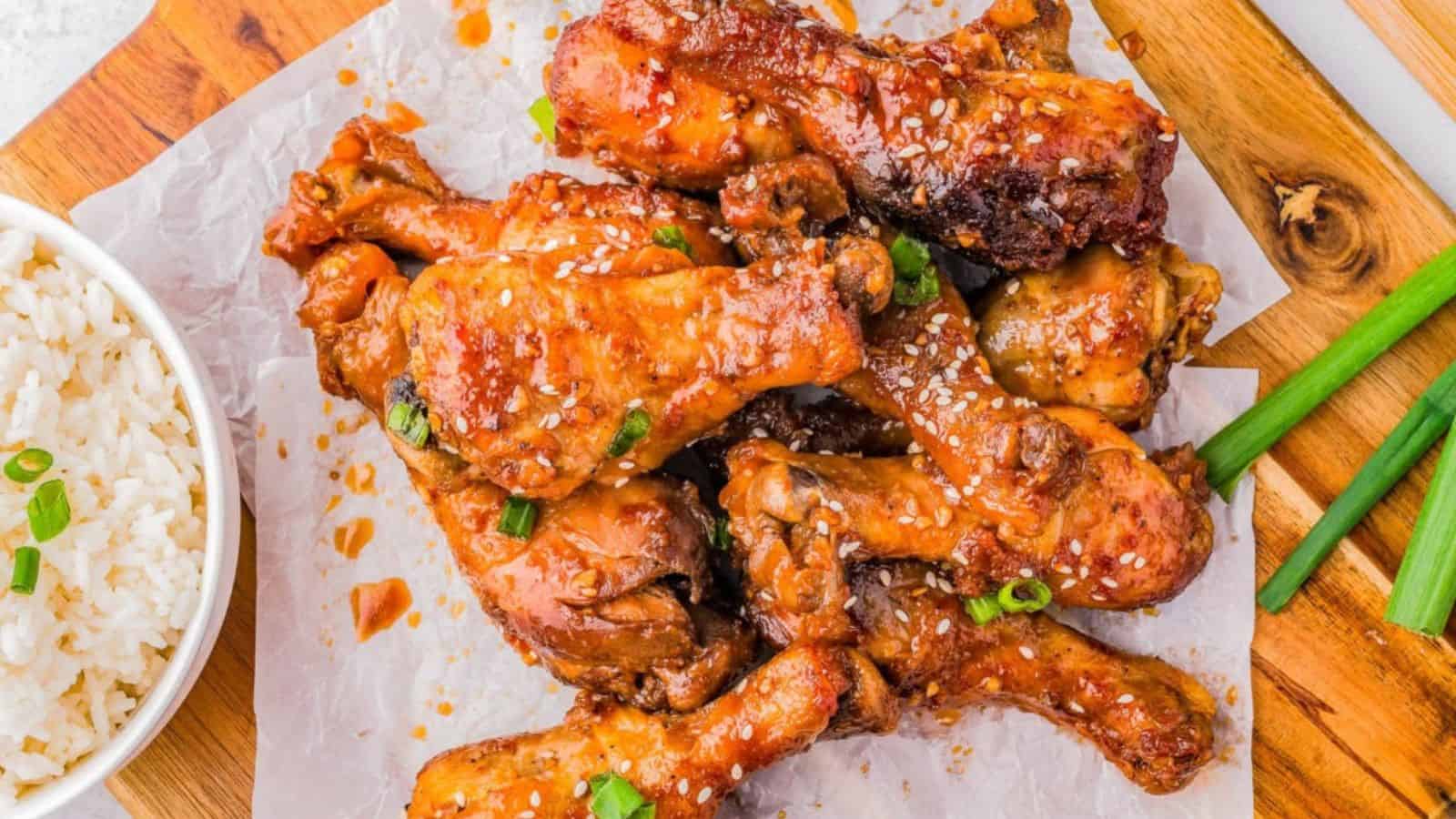 A platter of glistening, sesame-coated chicken drumsticks garnished with green onions, alongside a bowl of fluffy white rice.