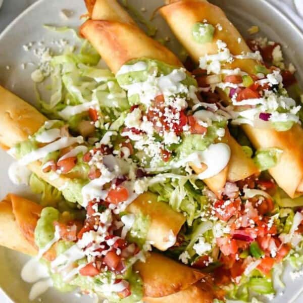 A plate of rolled tacos topped with shredded lettuce, diced tomatoes, guacamole, cheese, and a dollop of sour cream.
