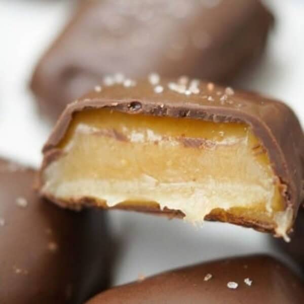 A close-up of a chocolate-covered candy with a bite taken out reveals layers of caramel and creamy filling, sprinkled with sea salt.