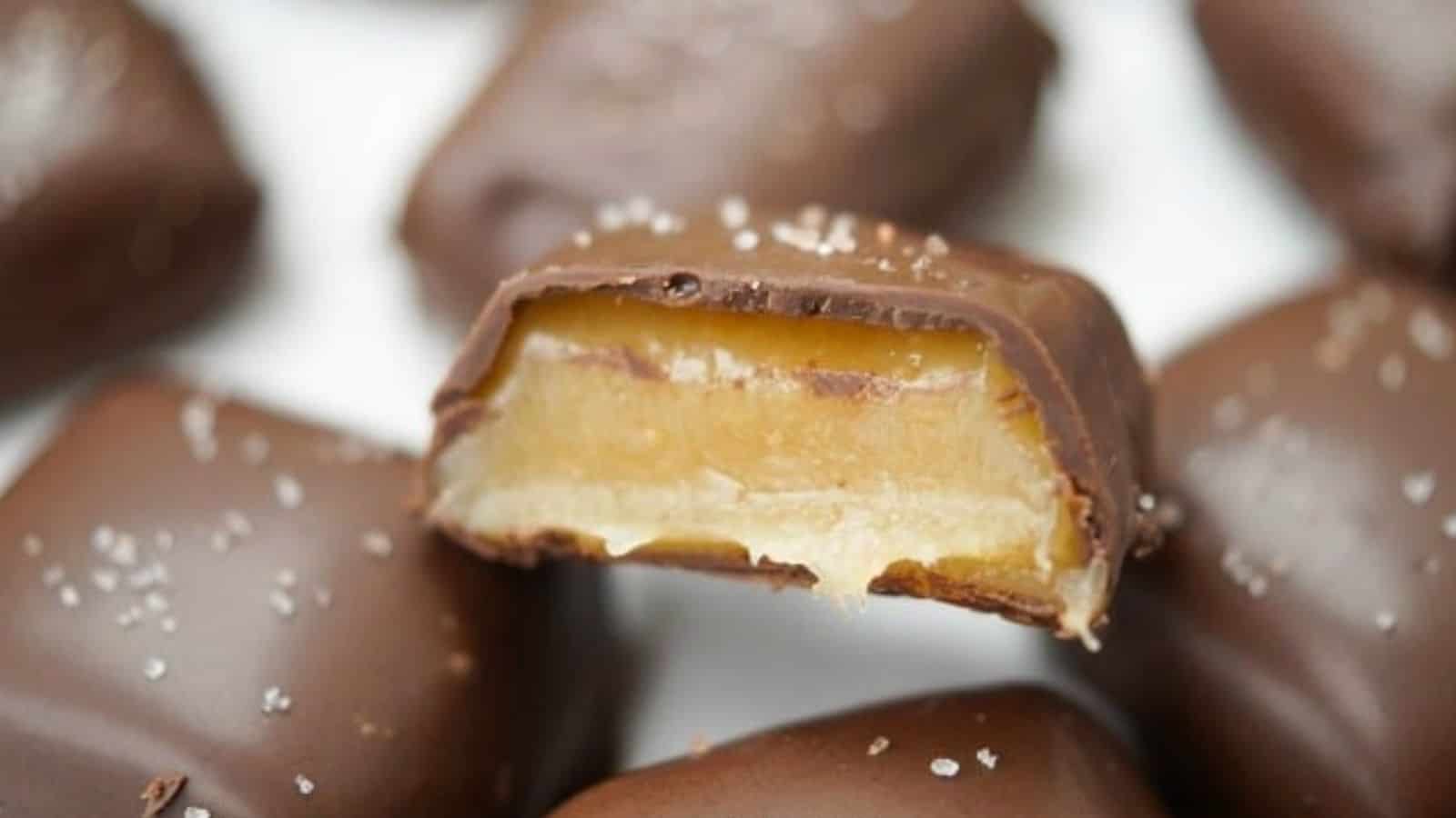 A close-up of a chocolate-covered dessert with a bitten piece revealing a creamy caramel filling, topped with a sprinkle of salt.