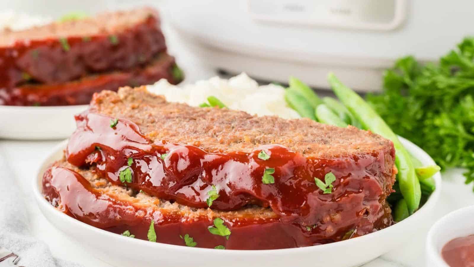 A close-up of a slice of meatloaf topped with glossy ketchup glaze, garnished with parsley, served with green beans and mashed potatoes.