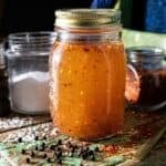 A jar of orange sauce is on a rustic chair, accompanied by a pepper grinder, salt, and an extra jar of chili flakes.