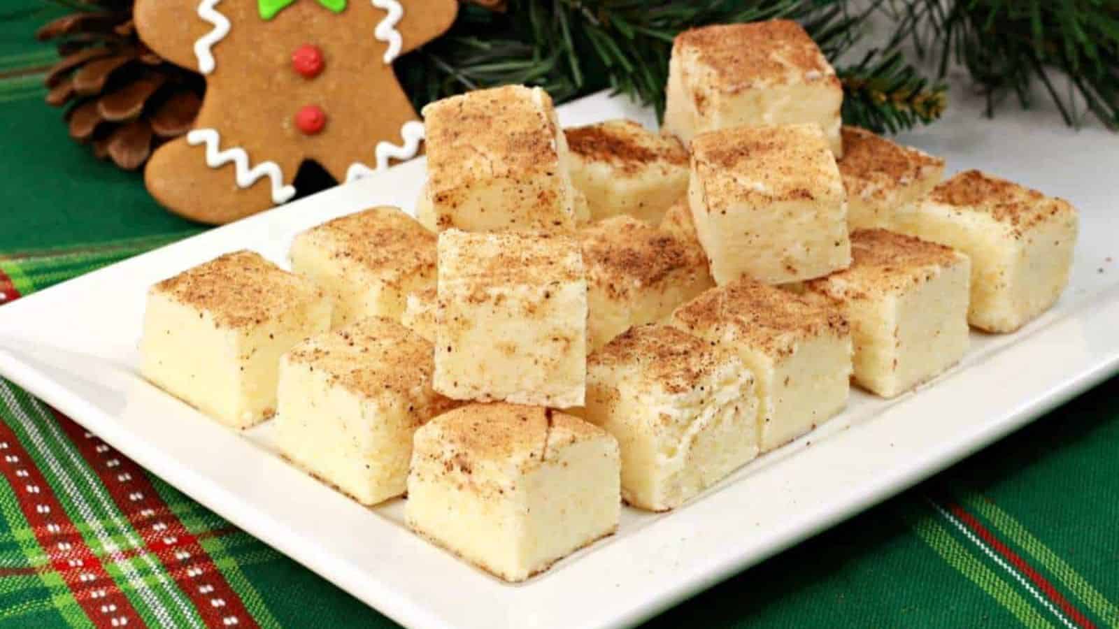 A white plate holds square pieces of creamy fudge sprinkled with cinnamon, set against a festive green tablecloth with pine branches.
