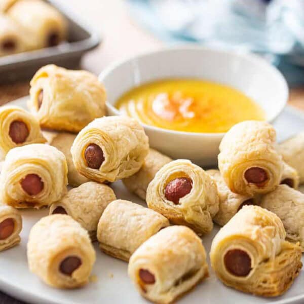 A plate of pig in a blanket appetizers with flaky pastry surrounding small sausages, accompanied by a bowl of mustard dipping sauce.