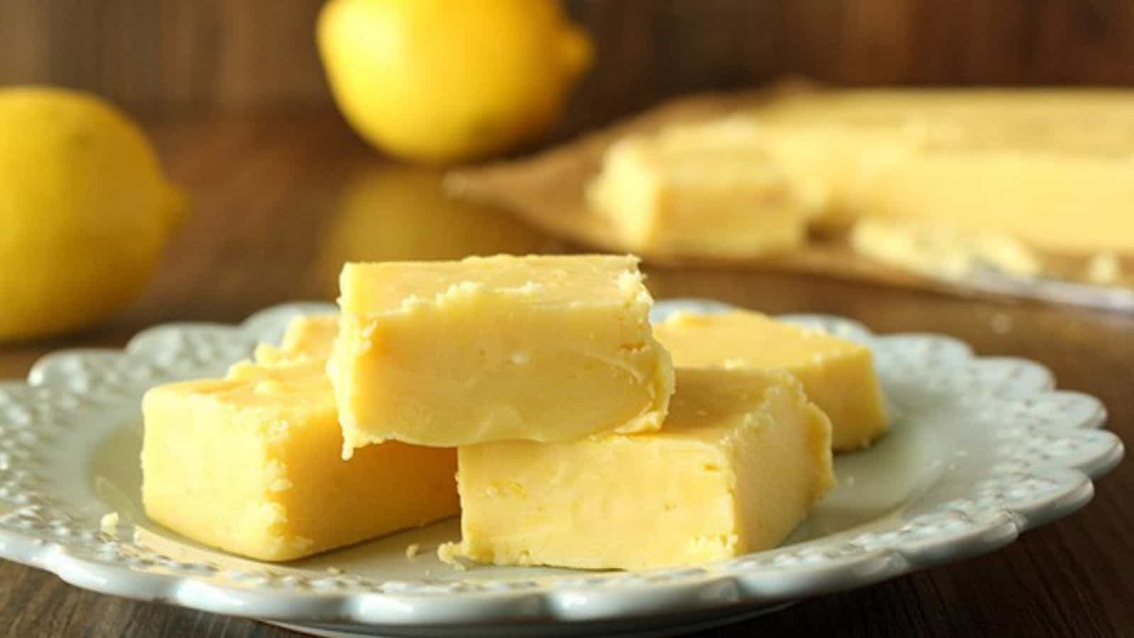Three pieces of yellow lemon fudge on a decorative white plate, with fresh lemons in the background.
