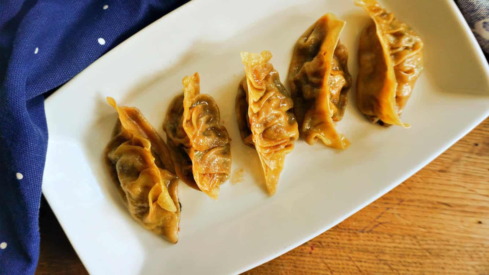 Five cooked dumplings are arranged in a row on a white rectangular plate. The plate is placed on a wooden surface, and a dark blue fabric with white dots is partially visible in the top left corner.