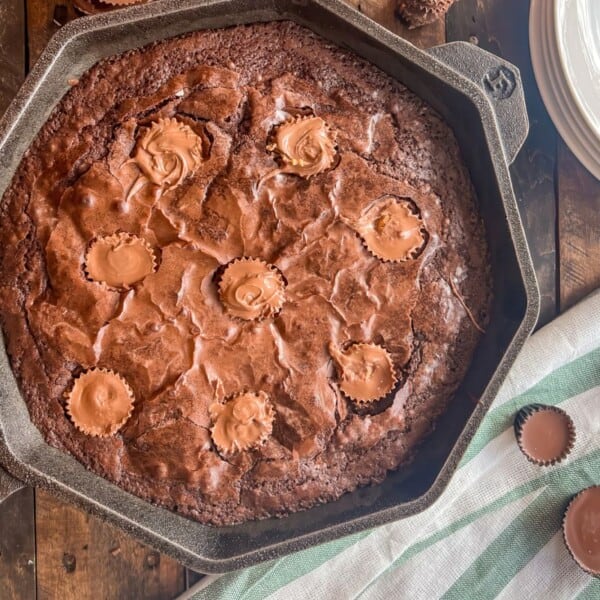 A skillet with a freshly baked brownie topped with peanut butter cups rests on a wooden surface. A white and green striped cloth is nearby, along with a stack of white plates on the side.
