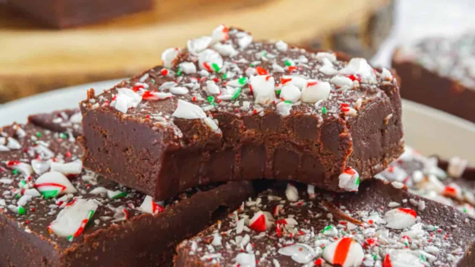A close-up of creamy chocolate fudge topped with crushed peppermint and festive green and red sprinkles, stacked on a plate.