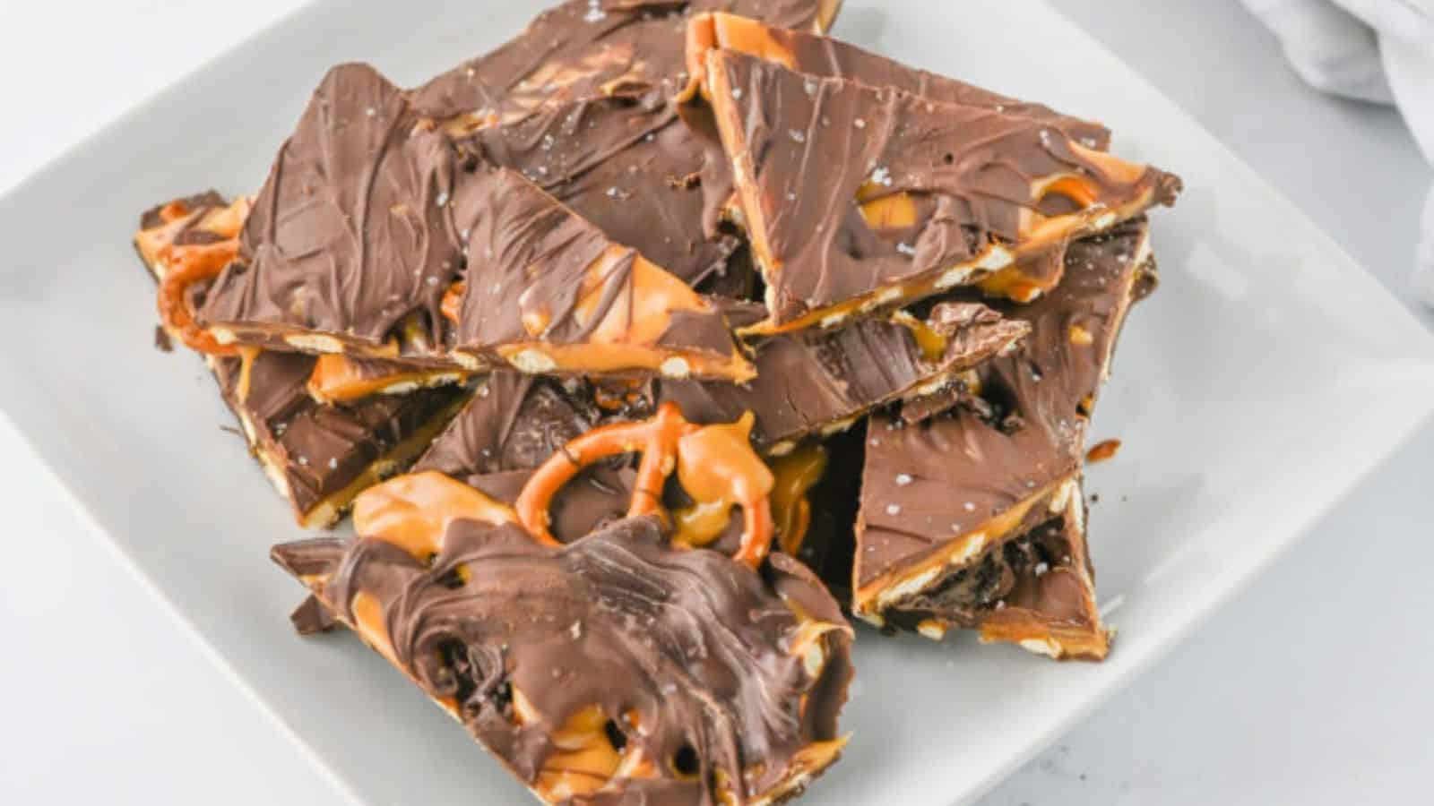 A plate stacked with chocolate-covered caramel pretzel bark, cut into triangular pieces and sprinkled with sea salt.
