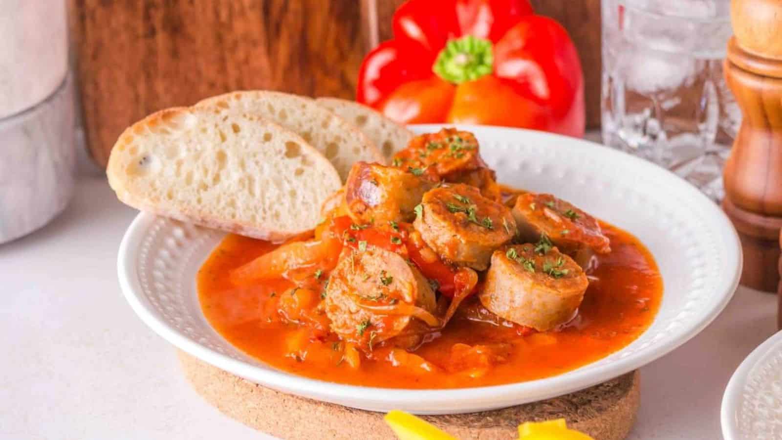 A plate of sausage and bell pepper stew garnished with herbs, accompanied by slices of rustic bread and a red bell pepper in the background.