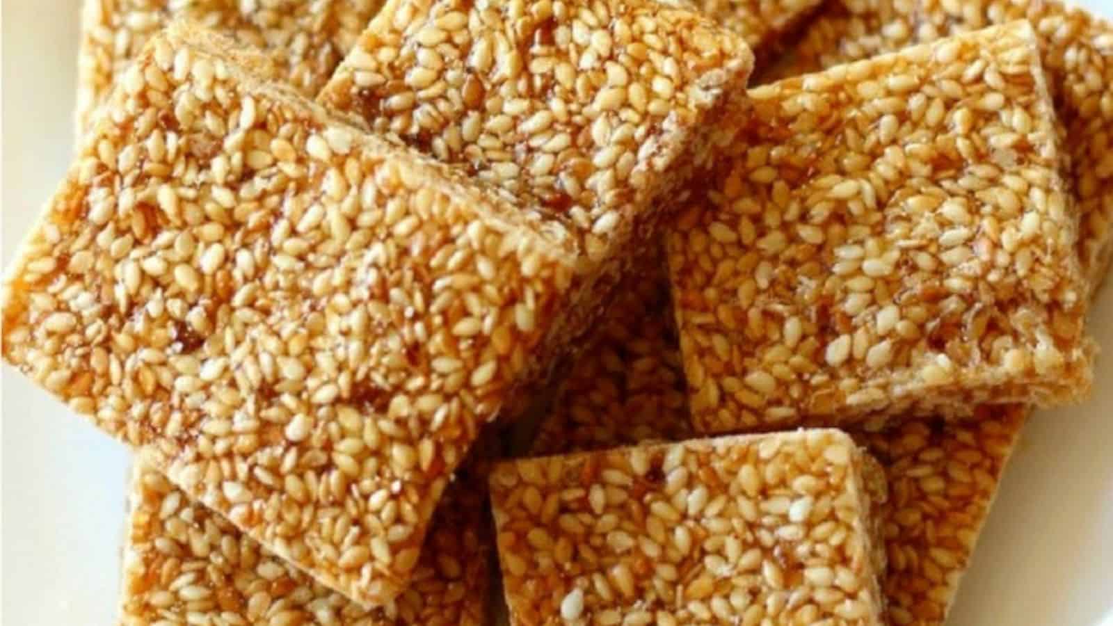 A close-up of square sesame candy pieces, golden-brown and covered in sesame seeds, arranged on a white plate.