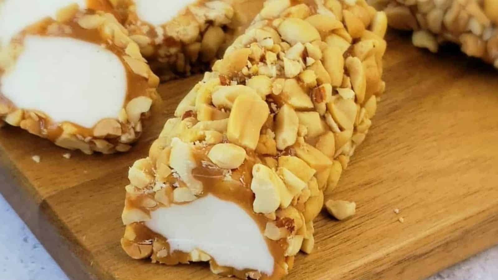 Close-up of a creamy, white dessert coated in caramel and crushed peanuts, sliced on a wooden board. Nutty and sweet treat.