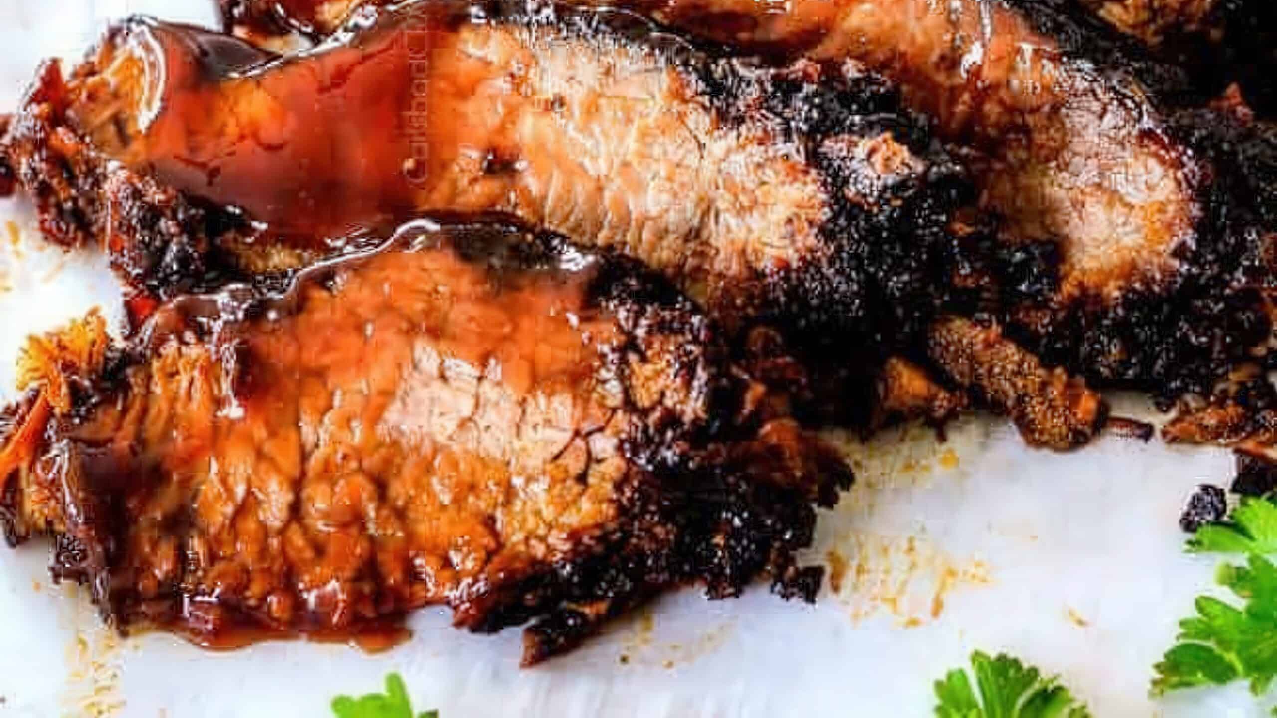 Close-up of several slices of glazed barbecued meat, arranged on a white surface. The meat appears juicy and caramelized with charred edges. Fresh parsley leaves are scattered nearby for garnish.
