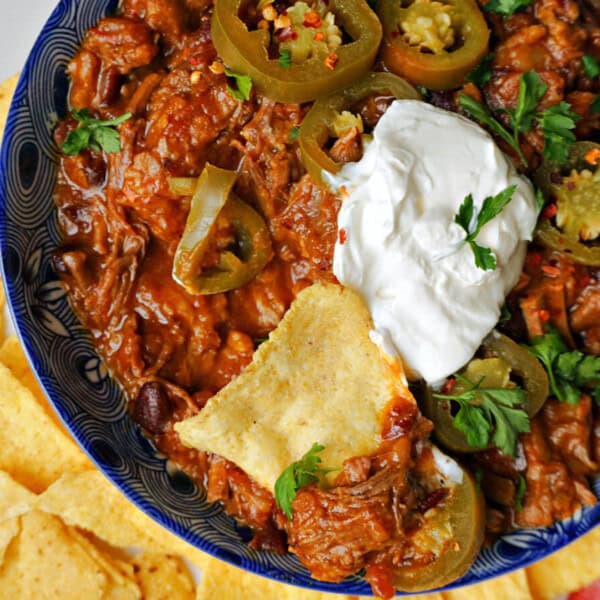 A bowl of chili topped with sour cream, jalapeños, and herbs is served on a plate with tortilla chips surrounding it.