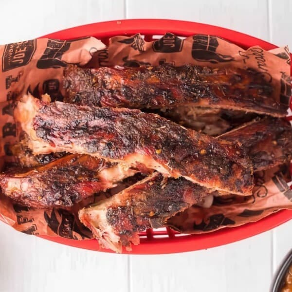 Barbecue ribs in a red plastic basket lined with branded paper, placed on a white surface. Sides of coleslaw and baked beans are partially visible around the basket.