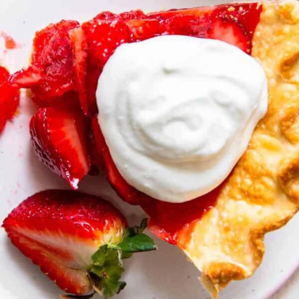 A slice of strawberry pie topped with whipped cream, accompanied by a fork and fresh strawberry slices, served on a white plate.