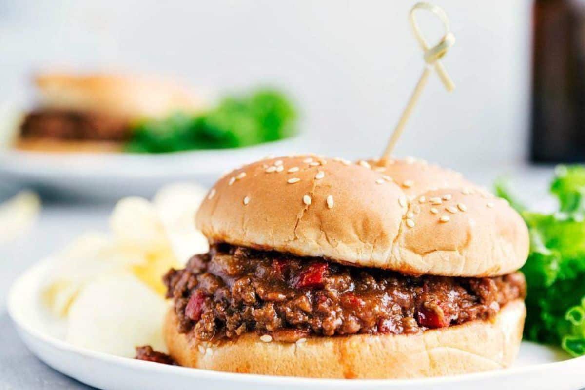A close-up of a Sloppy Joe sandwich on a sesame seed bun, served on a white plate with potato chips and a garnish of lettuce in the background. A cocktail stick holds the sandwich together.