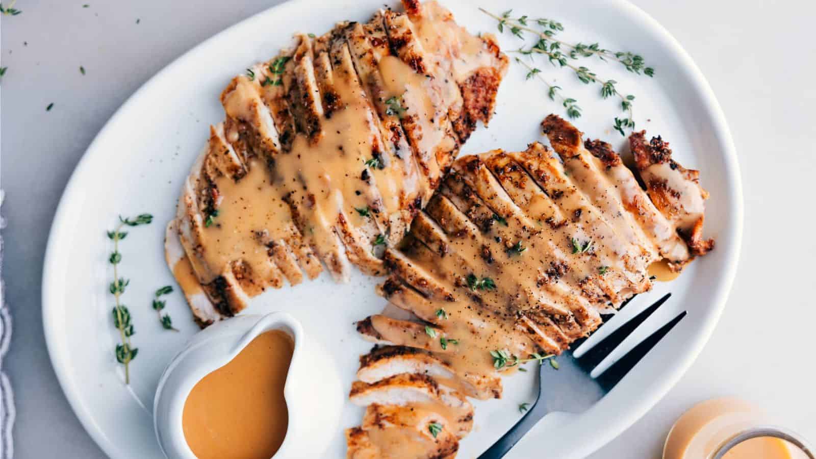 Sliced grilled chicken breast drizzled with sauce, garnished with thyme, served on a white plate with a small sauce bowl.