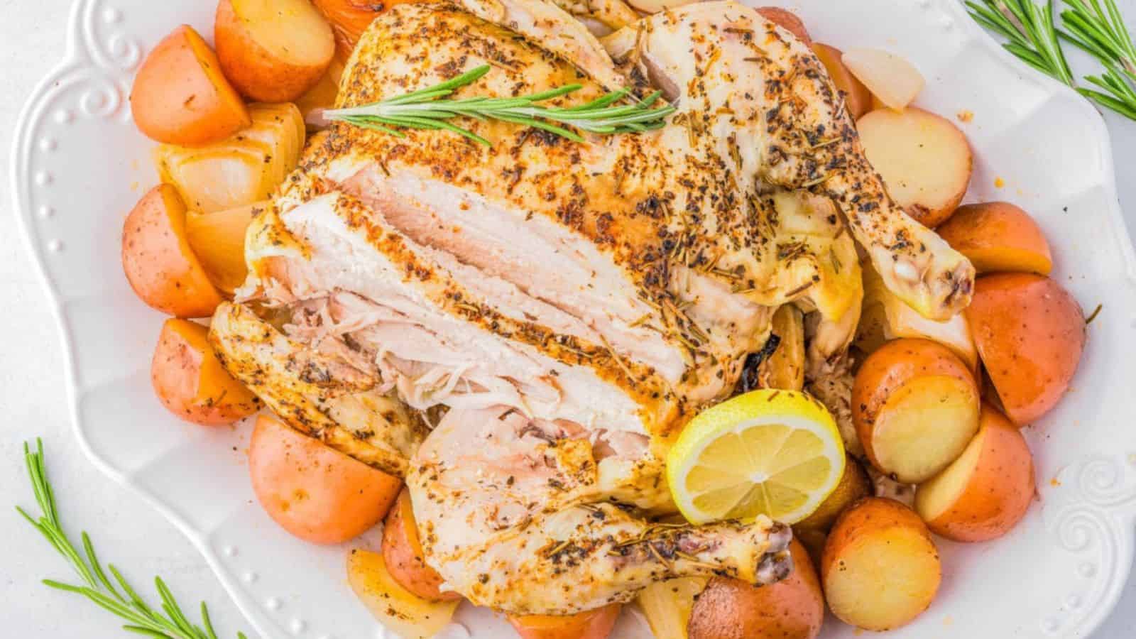 A roasted chicken, sliced and garnished with lemon and rosemary, served on a white platter with roasted red potatoes surrounding it.