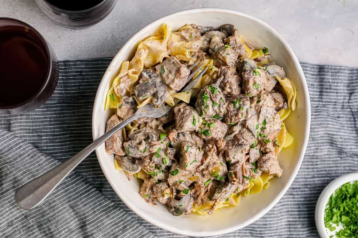 A bowl of beef stroganoff with creamy sauce, mushrooms, and noodles, garnished with chopped parsley. A fork rests on the side of the bowl. Nearby are a glass of red wine and a small dish of green herbs on a gray-striped cloth.