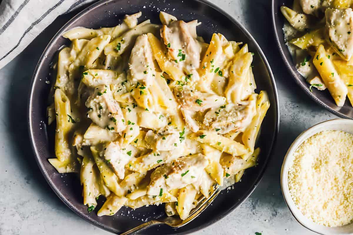 A plate of creamy chicken Alfredo pasta garnished with chopped parsley. The dish features penne pasta and sliced chicken pieces in a rich, white sauce. A small bowl of grated Parmesan cheese is beside the plate.