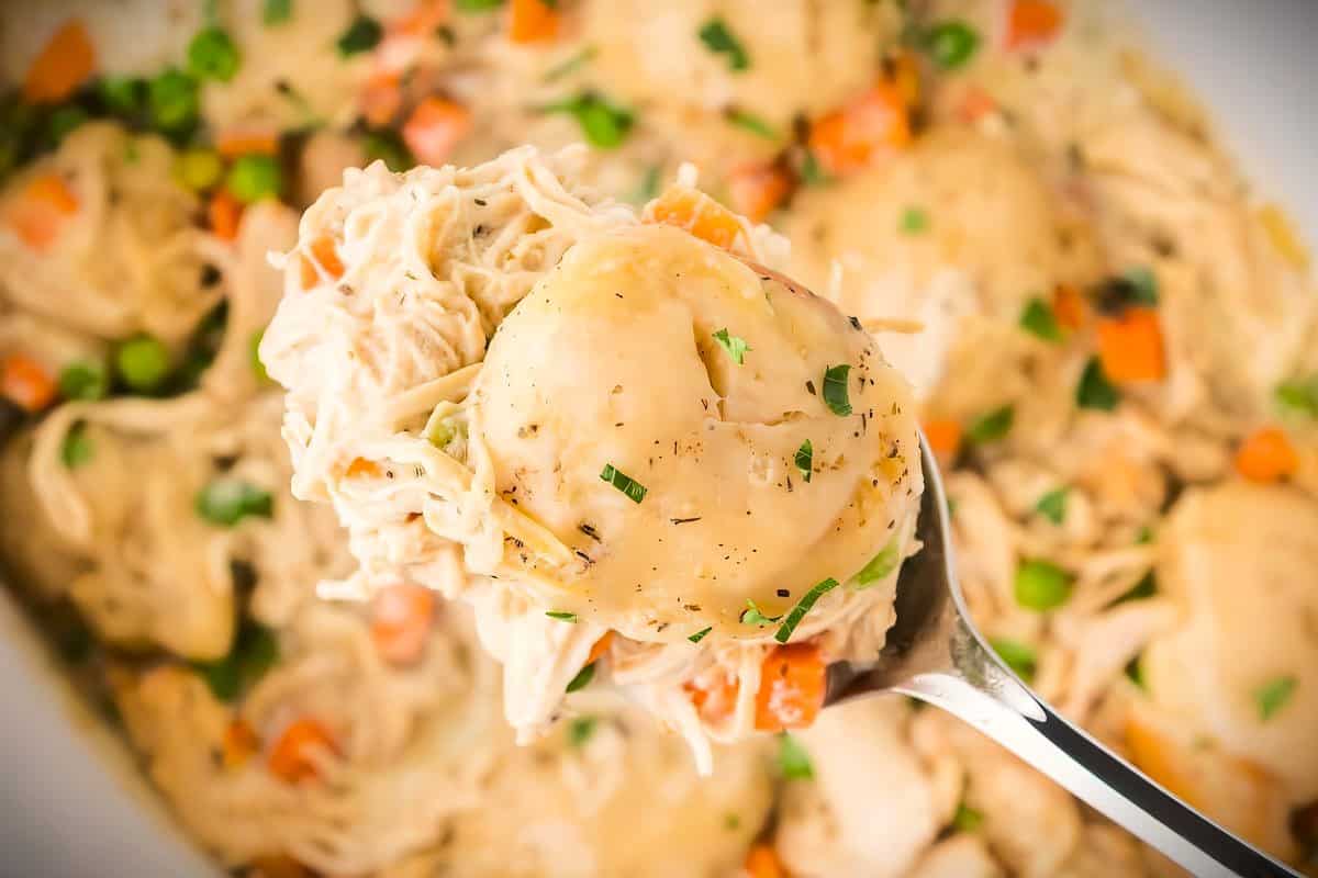 A close-up of a spoon lifting a portion of chicken and dumplings from a pot. The dumplings are creamy and surrounded by shredded chicken, peas, and diced carrots seasoned with herbs. The dish has a comforting, hearty appearance.