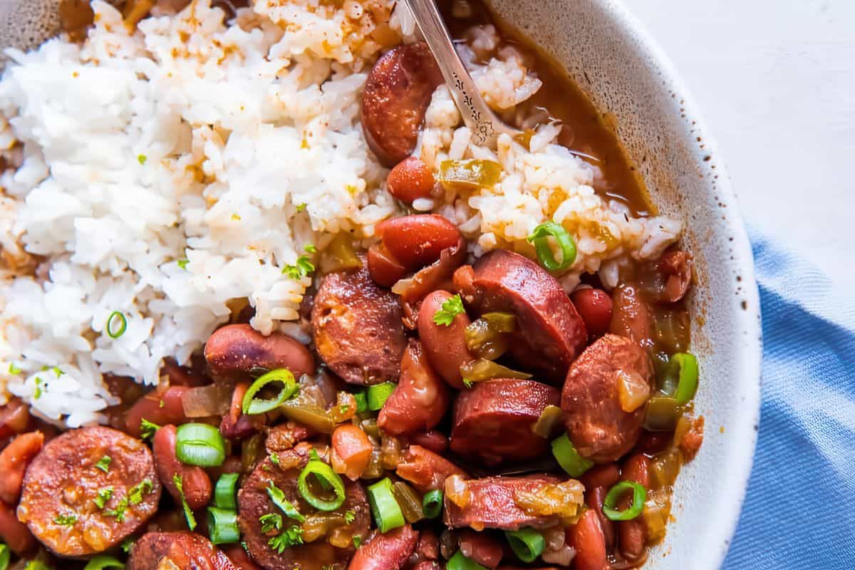 A bowl split with cooked white rice and a red bean stew. The stew contains sliced sausage, red beans, and chopped green onions. A spoon is tucked into the dish, and a blue cloth is partially visible underneath the bowl.