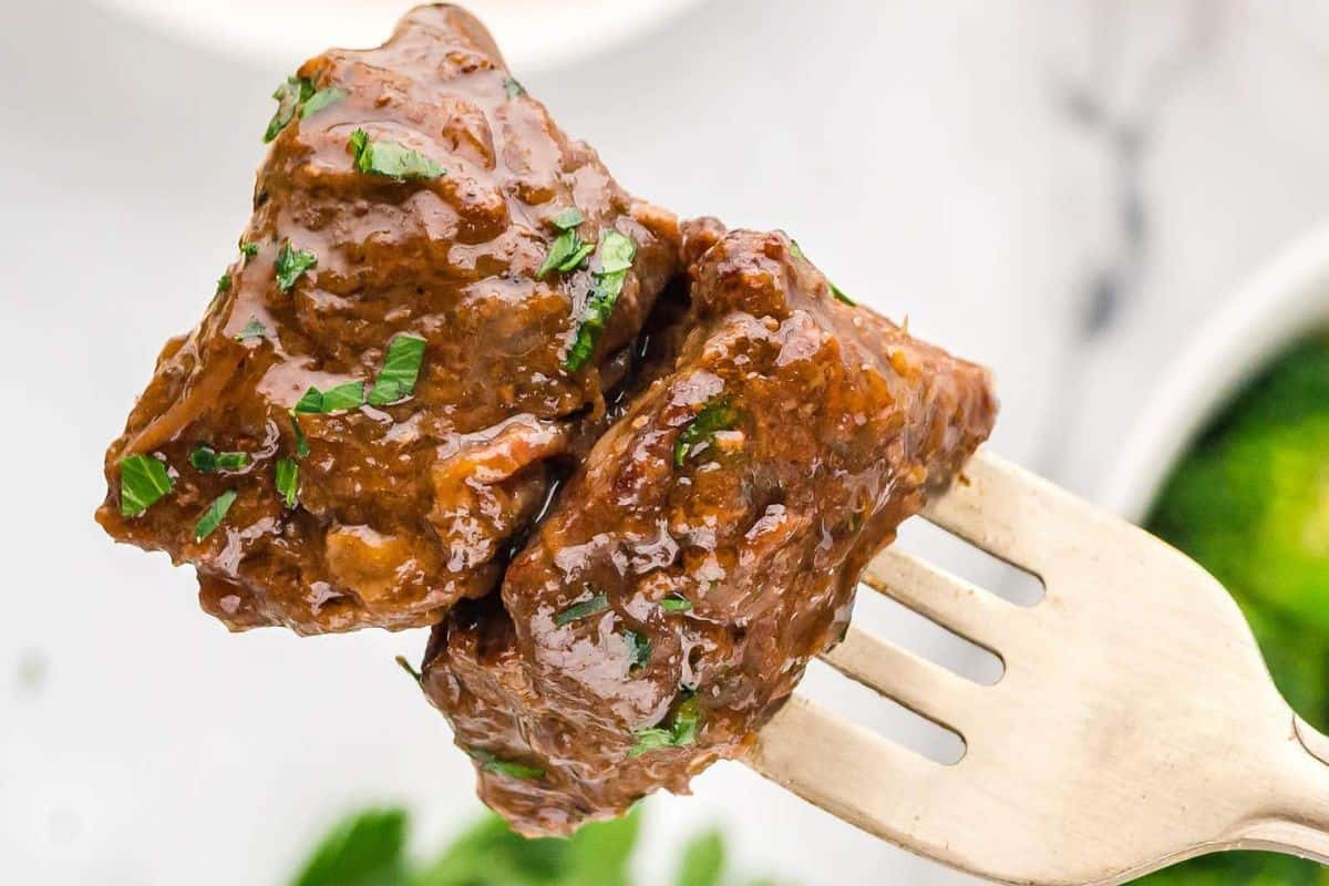 Close-up of a fork holding two pieces of braised beef covered in a rich, glossy sauce, garnished with chopped herbs. The background is softly blurred, with hints of a green vegetable, possibly broccoli, visible.