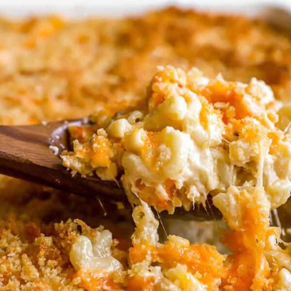 A close-up shot of a wooden spoon scooping out a serving from a baking dish of baked macaroni and cheese. The dish features a golden-brown breadcrumb topping over creamy, melted cheese mixed with macaroni pasta.
