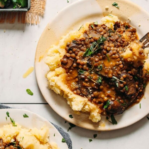 A plate of mashed potatoes with lentil stew and herb garnish, and a hand holding a fork positioned above the food.