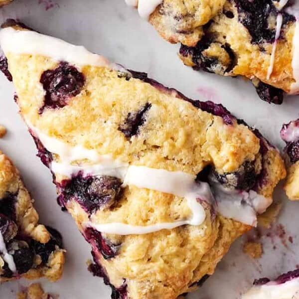 Close-up of blueberry scones on a white surface, each drizzled with white icing. The scones are golden brown with visible blueberries, and some have a sugared appearance.