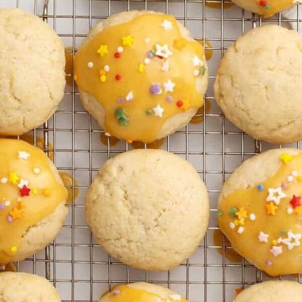 The image features nine cookies resting on a wire rack. Some cookies are decorated with orange icing and colorful star-shaped sprinkles. A white striped cloth is positioned next to the rack.