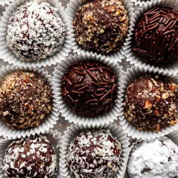 An assortment of chocolate truffles in paper cups, topped with powdered sugar, nuts, shredded coconut, and chocolate sprinkles, arranged in a grid pattern.