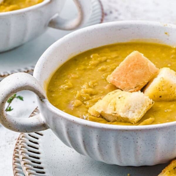 A bowl of pea soup with croutons on top is placed on a white plate, accompanied by a sprig of thyme. Additional bowls are visible in the background.