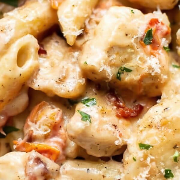 A close-up view of pasta with a creamy sauce, chunks of chicken, herbs, and small pieces of tomato in a white bowl.