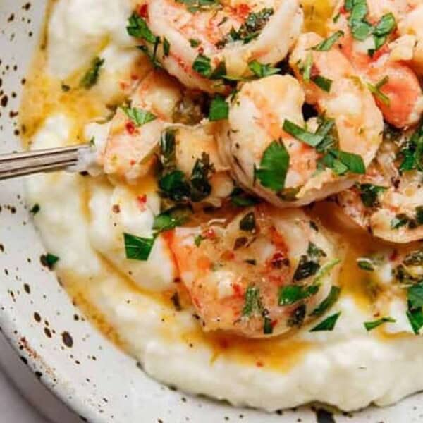 A speckled bowl containing shrimp in a garlic butter sauce sits on top of creamy mashed potatoes, garnished with chopped parsley. A fork is resting on the side of the bowl.