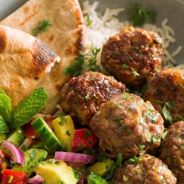 The image features a plate with meatballs on rice, accompanied by pita bread, a cucumber-tomato salad, and a mint garnish.