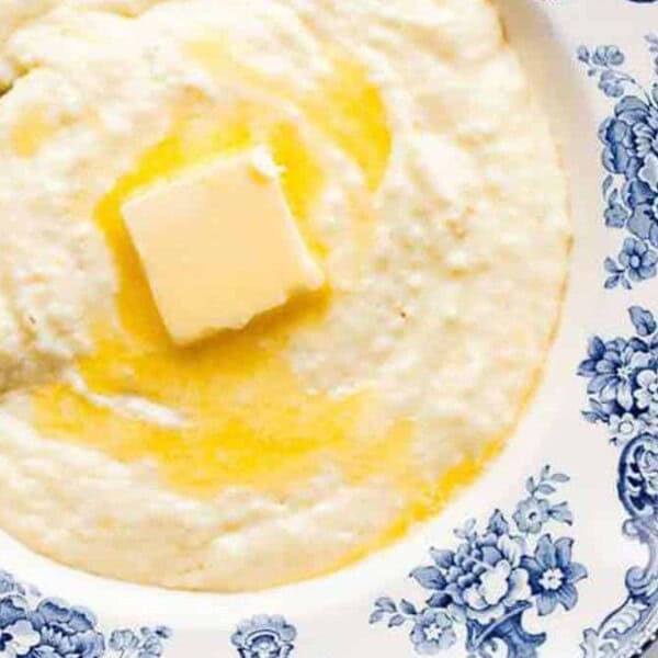 A blue bowl with a floral pattern containing creamy grits topped with a pat of melting butter, accompanied by a spoon on the side.