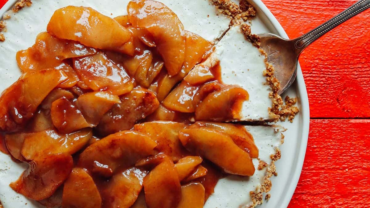 A pie with a cream base, topped with glazed apple slices and partially cut, sits on a plate. A spoon rests on the pie. The background features a red wooden surface.