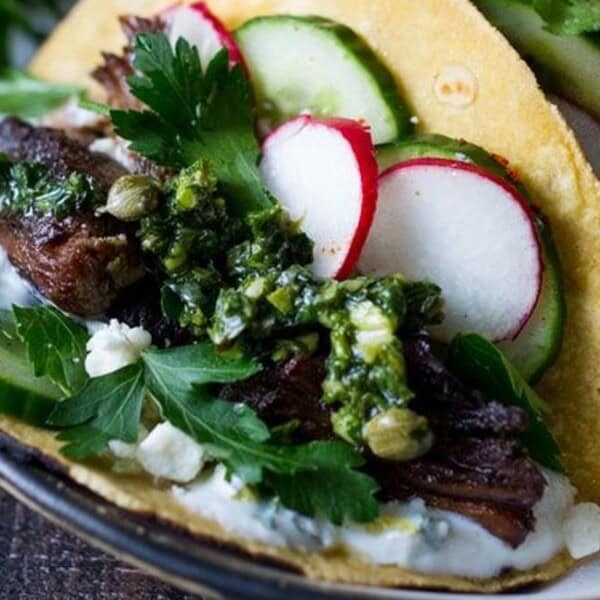 Tacos filled with grilled meat, sliced cucumbers, radishes, fresh parsley, and a dollop of sauce on a tortilla. Garnished with a green herb mixture, served on a round plate.