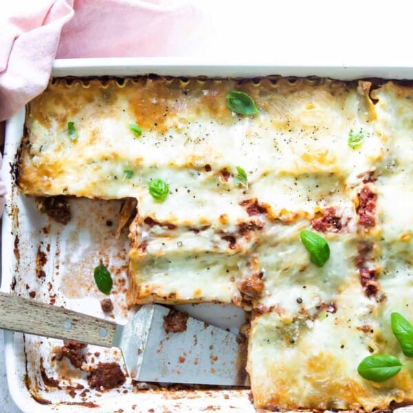 A white dish containing lasagna with a golden brown top, garnished with basil leaves. A spatula is placed in an area where a portion has been removed.