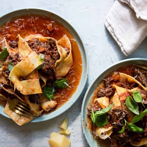The image depicts two plates of pasta topped with a rich meat sauce and garnished with fresh basil. They are placed on a textured surface, alongside shards of Parmesan cheese, napkins, and forks.