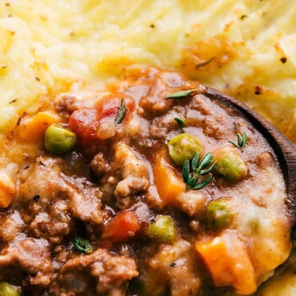 The image shows an autumn pie topped with herbs, featuring layers of minced meat, peas, carrots, and corn beneath creamy mashed potatoes. A wooden spoon is ready to scoop the pie.