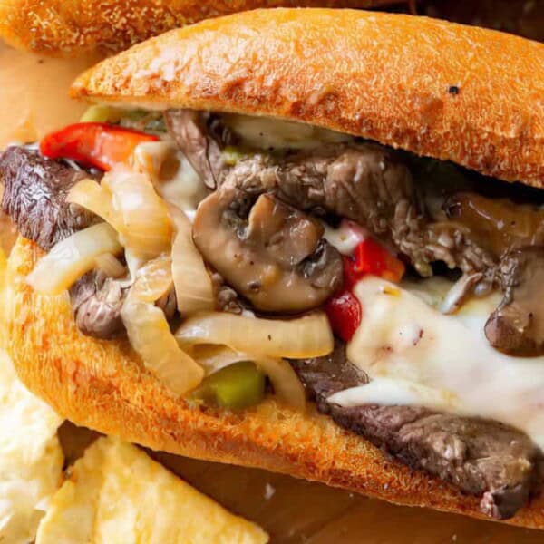 A close-up of a Philly cheesesteak sandwich with sliced beef, melted cheese, mushrooms, green peppers, and caramelized onions in a crusty roll. Pieces of flatbread are scattered around on a wooden surface.