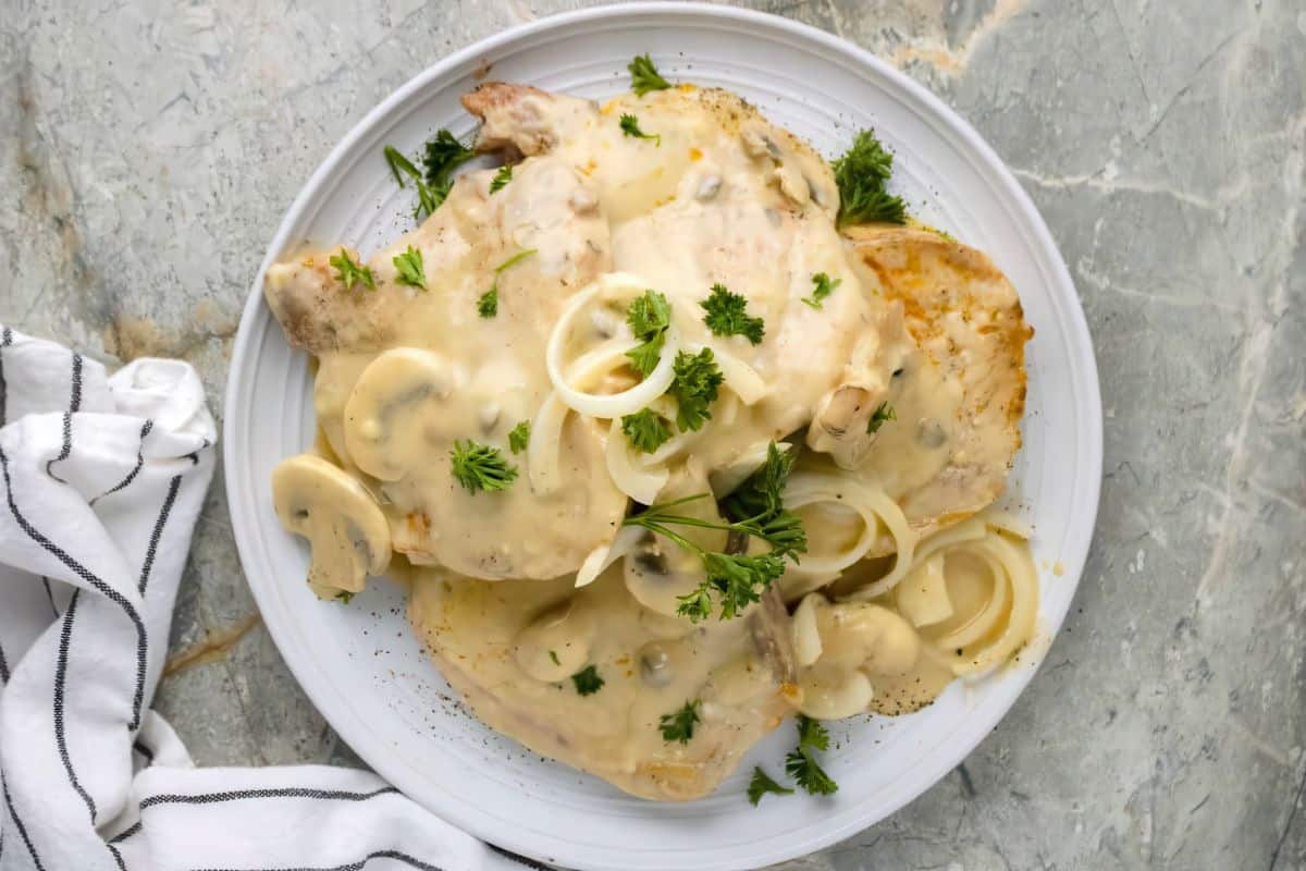 Plate of pork chops covered in creamy mushroom sauce, garnished with sliced onions and fresh parsley. A striped cloth napkin is partially visible on a gray marble surface.