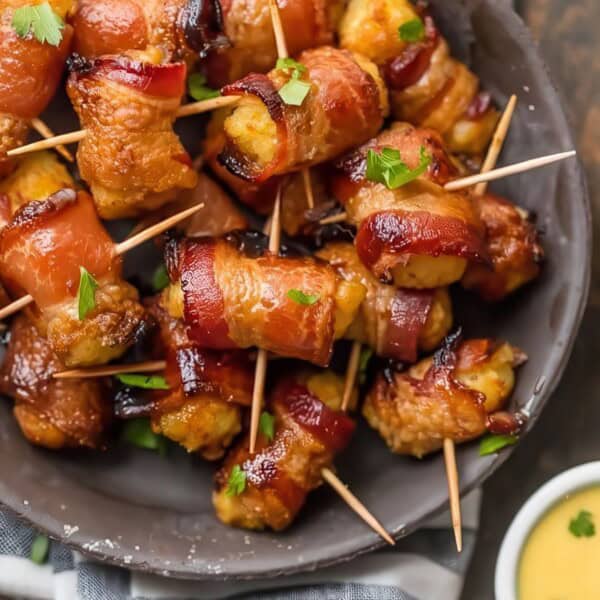 A bowl containing bacon-wrapped pineapple bites with toothpicks, garnished with parsley, next to small bowls of ketchup and mustard dipping sauces.