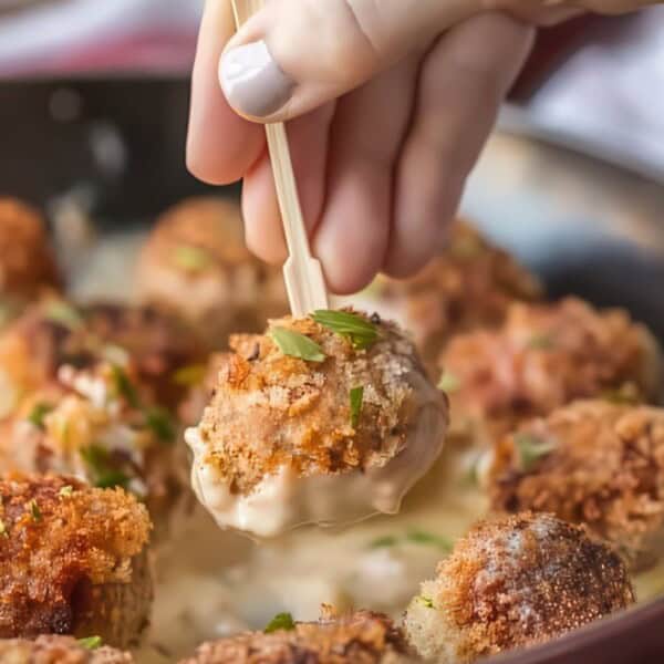 A hand holds a wooden pick, lifting a breaded meatball from a skillet filled with creamy sauce and topped with chopped herbs.